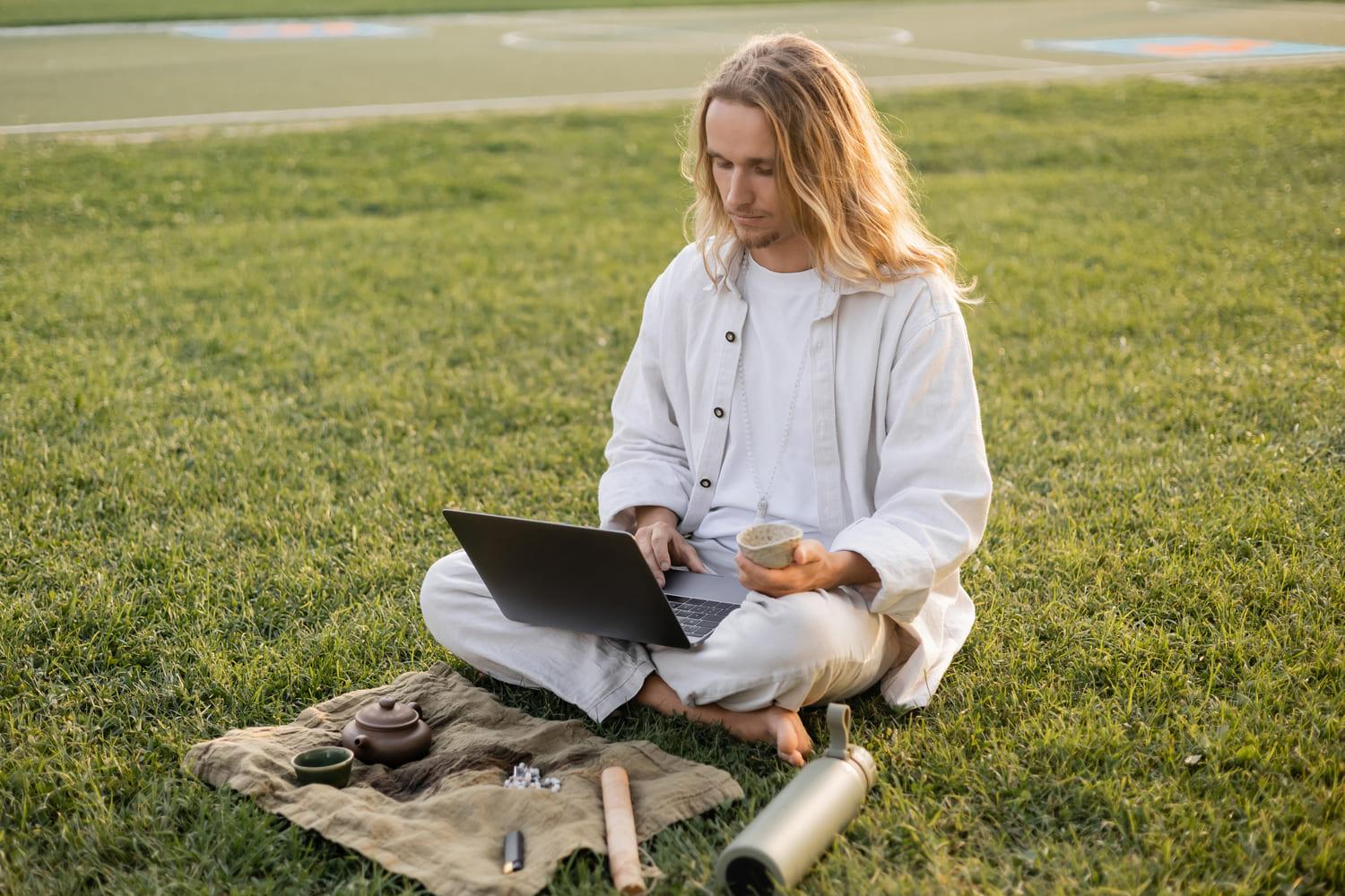 Spiritual Reading - Young birded man sitting on grass in white clothes holding tea in front of laptop